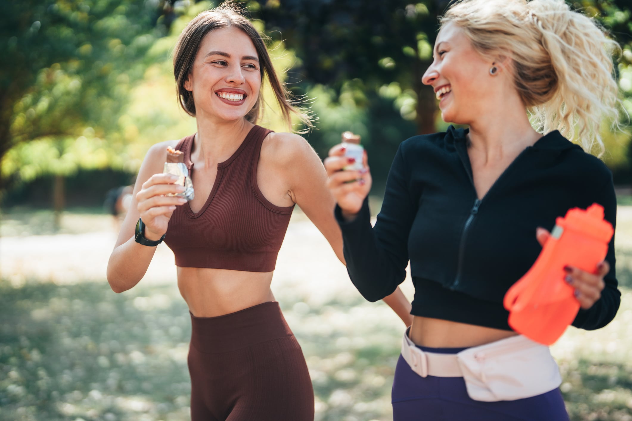 Two fit friends walking together in a park after exercising, smiling and enjoying protein healthy bars. The scene captures post-workout camaraderie and the importance of nutrition and friendship in fitness routines.