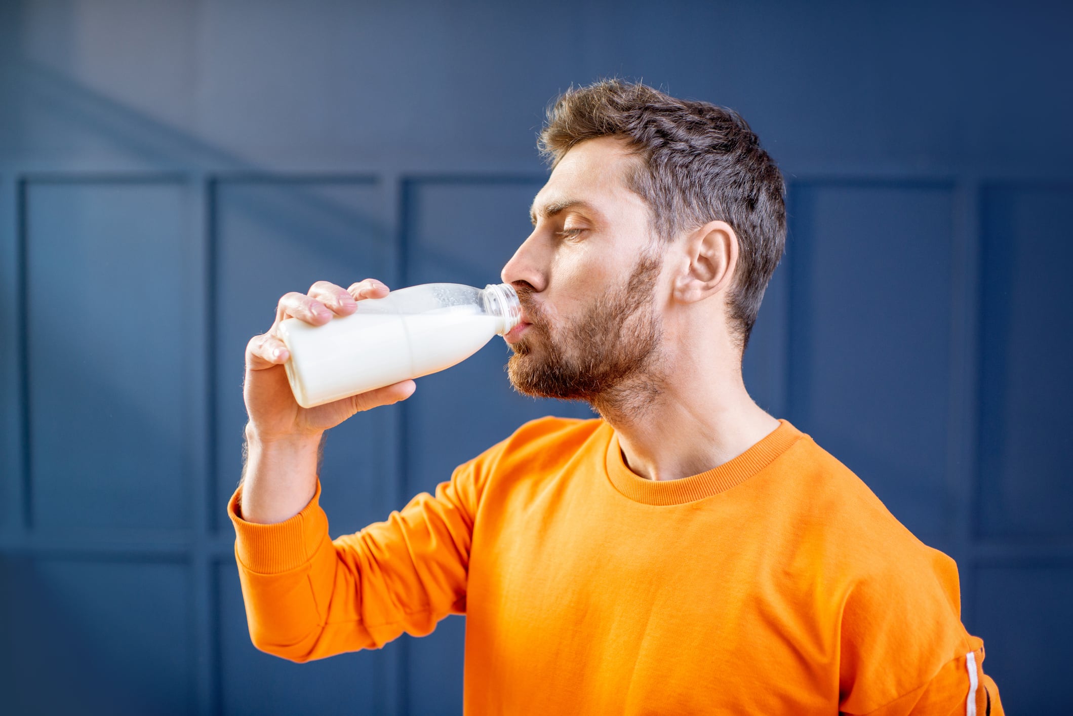 Conceptual portrait of a sports man in bright sweater drinking milk from the bottle on the blue background