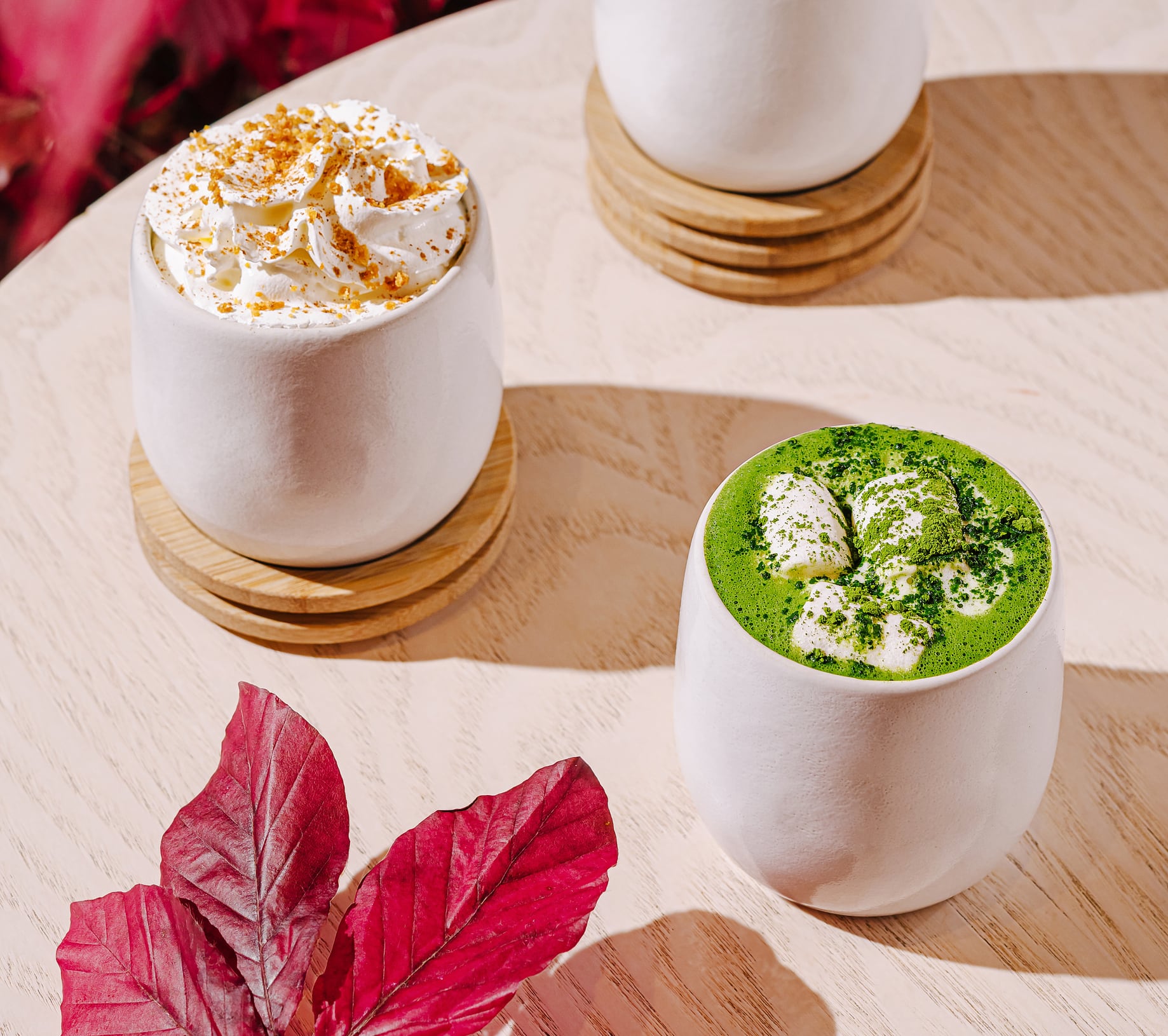 Two unique dessert beverages sit on a wooden table surrounded by vibrant leaves. One has green matcha with cream, while the other features whipped topping and spices.