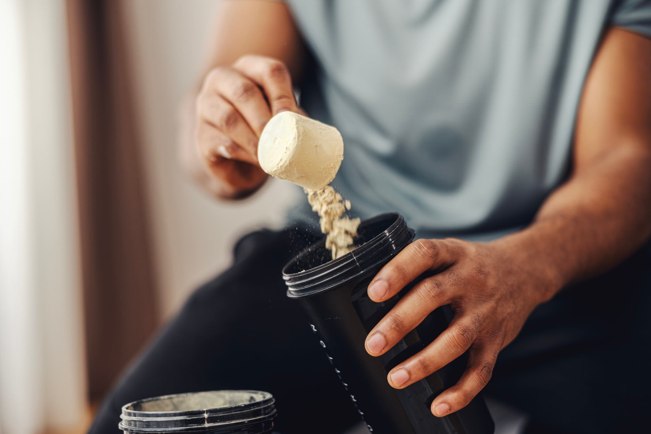 Adult man's hands scooping protein powder from a container into a black shaker bottle, preparing a nutritional supplement drink for workout recovery and muscle building