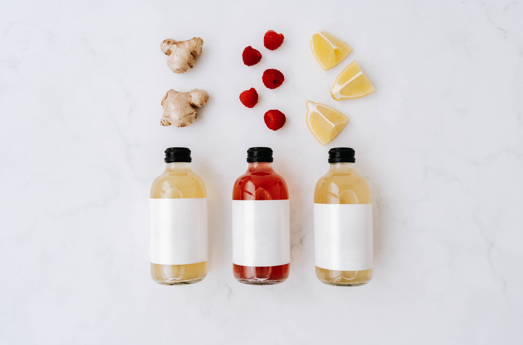 Three small transparent glass bottles with black caps, filled with colourful kombucha of yellow, red, and green and ingredients on a white surface. Each bottle has a plain white label. Top view. Copy space