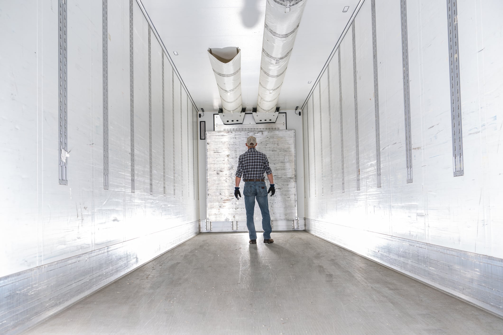 A worker in a checkered shirt and gloves stands in the middle of a refrigerated truck trailer. The interior is white and spacious, with visible cooling equipment on the ceiling. The worker appears focused as he inspects the area, ensuring it is clean and ready for cargo transport.