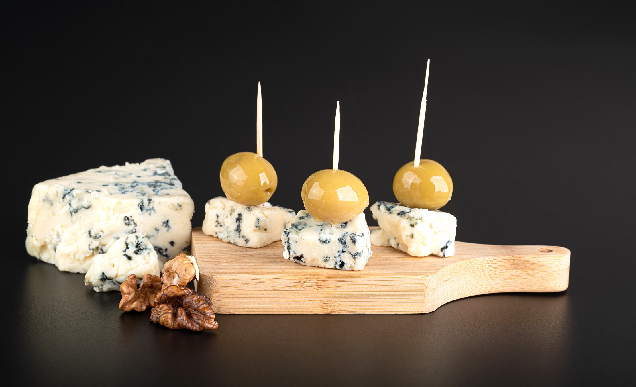 Cheese with blue mold and large green olives on a wooden cutting board, isolated on a black background with reflection petrova_dc