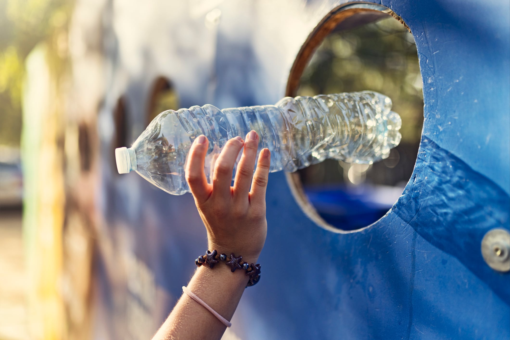 Close-up of teenage girl hand engage in responsible waste disposal at a public plastic recycling station. Teenage girl, dressed in a red shirt, is placing plastic bottles into a circular slot on a bold blue recycling wall marked with “PLÁSTICA.”
Shot with Canon R5