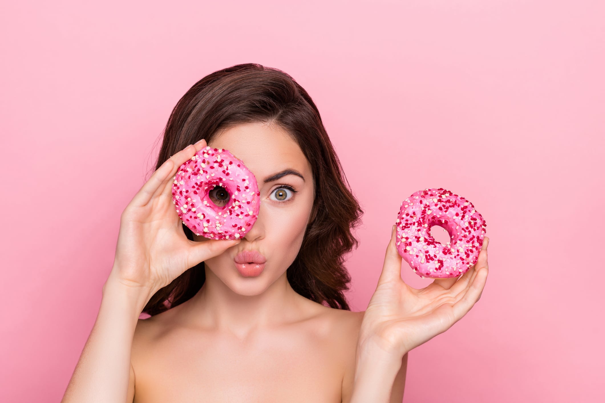 Woman holding two doughnuts - one over eye. Pink background.