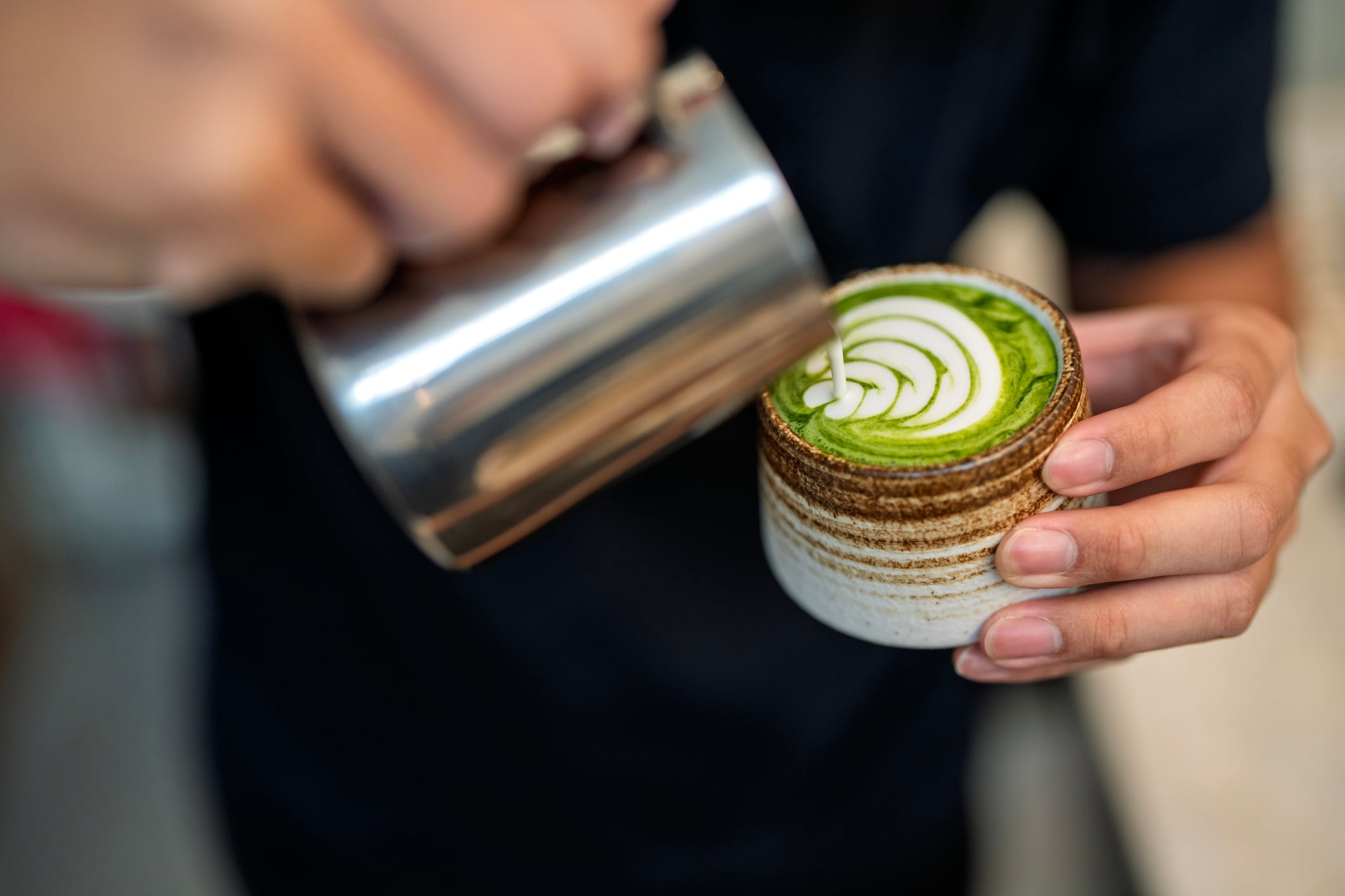 Close-up of a barista pouring steamed milk into a ceramic cup of matcha latte, creating detailed latte art during preparation.