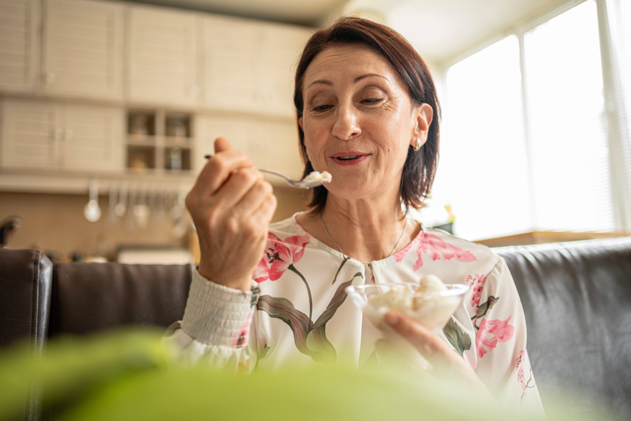 Smiling mature woman savoring a sweet frozen treat, eyes closed for pleasure, eating ice cream with a spoon in a bright cozy indoor setting