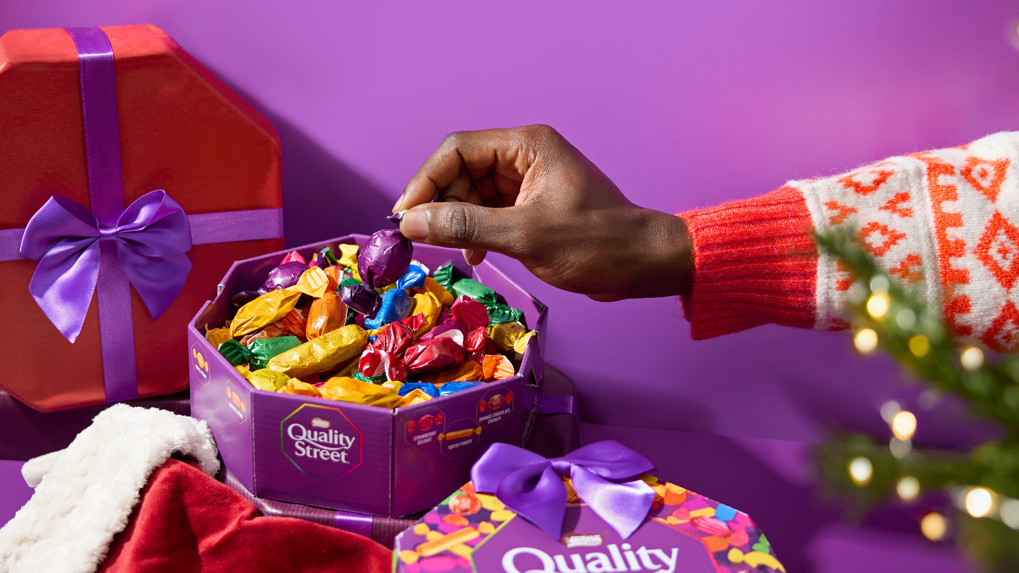 Tub of Quality Street sweets with a hand reaching in and pulling out one of the sweets.