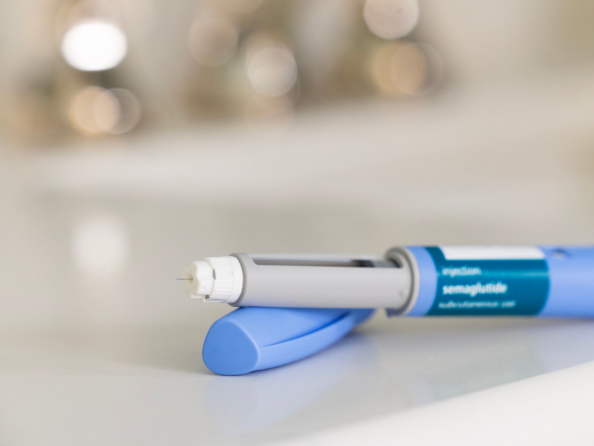 GLP-1 semaglutide injection pen, a medication used to treat Type 2 diabetes and aid in weight loss, on a white surface. The prescription medicine is used to treat Type 2 diabetes as It helps regulate blood sugar levels by stimulating insulin secretion and decreasing glucagon levels. In addition, it is also used off-label for weight loss in individuals with obesity, as it can help reduce appetite and increase feelings of fullness. The pen is positioned horizontally, with its cap removed, on a bathroom countertop with a white surface. The medical device is in the foreground and the background is out of focus because of a shallow depth of field.