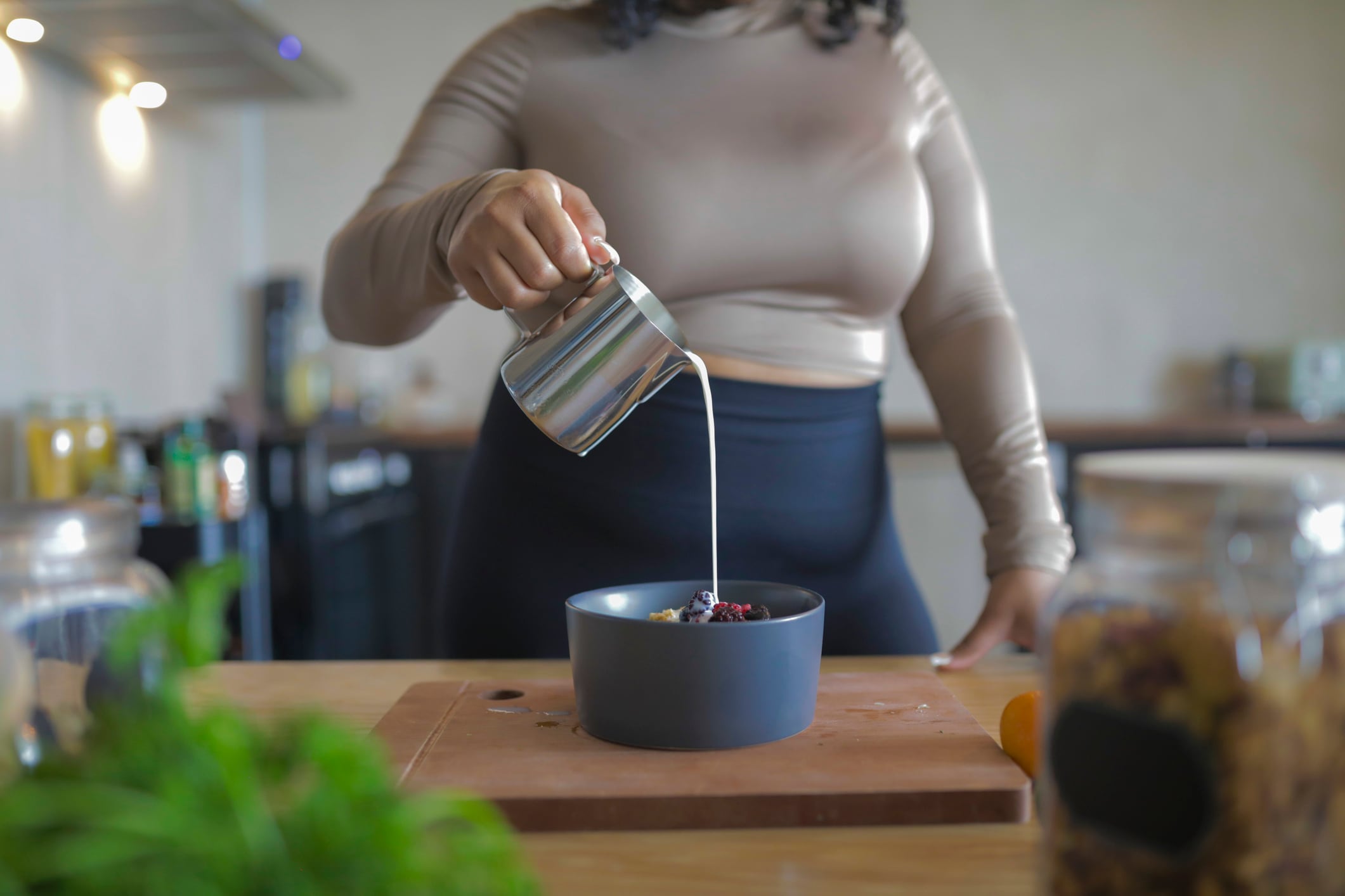 A woman pouring milk from a metal jug into a bowl of serial with fruit, in her kitchen at home.
